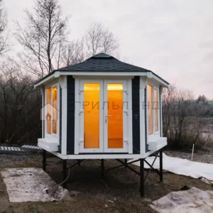 Всесезонная беседка с панорамными окнами «Гарден Хаус 9м²» на свайном фундаменте в скандинавском стиле на загородном участке при вечернем освещении.